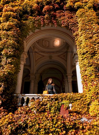 Young woman standing under arched column of an old building in autumn. Fall colors, foliage, one person, fashion, outfit, lifestyle.