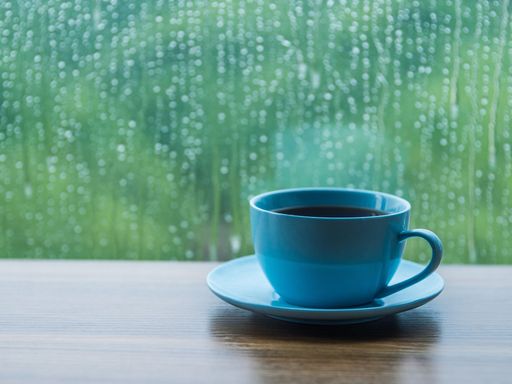 A blue cup of coffee on a wooden table in front of a window with water drops on a rainy day