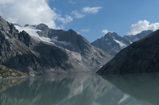 Calm lake water with a reflective surface surrounded by sunlit rough mountains under a beautiful cloudscape
