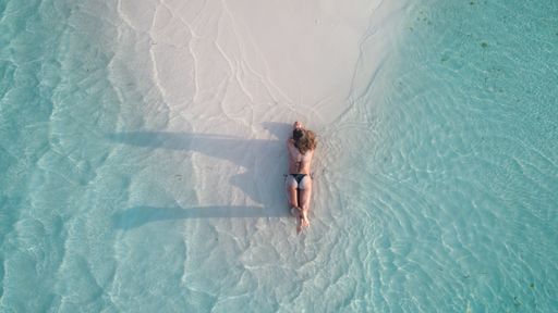 An aerial shot of a girl laying on the sand and tanning at the beach surrounded by clear blue water