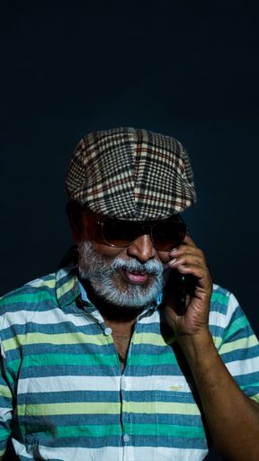 A portrait of a bearded Asian senior man wearing a hat and sunglasses talking on the phone isolated on a black background
