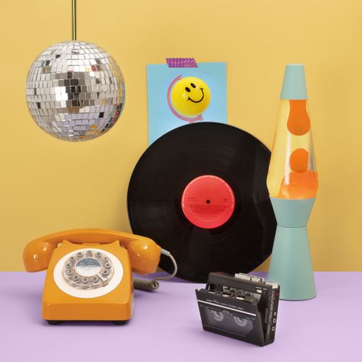 A ret cassette player, a lava lamp, an orange phone, and a vinyl disk on a purple table. Hanging on the yellow wall, there's a disco ball.