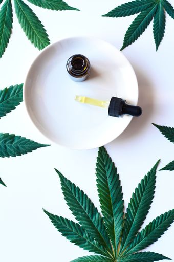 A top view of a serum bottle with a dropper on a plate with marihuana leaves on white surface