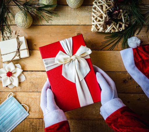 A closeup shot of hands in white gloves holding a Christmas present, decorations, and a sanitary mask on the table