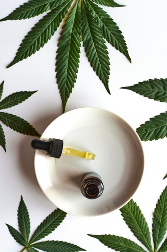 A top view of a serum bottle with a dropper on a plate with marihuana leaves on white surface