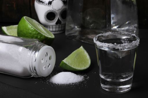 A tequila bottle and a salty jigger with lime and a Mexican skull next to them on a black surface
