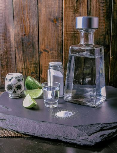 A tequila bottle and a salty jigger with lime and a Mexican skull next to them on a black surface