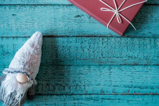 A top view of the gnome with a long, white beard and round nose in a gray hat near a red gift box on a blue, wooden surface - the concept of New Year