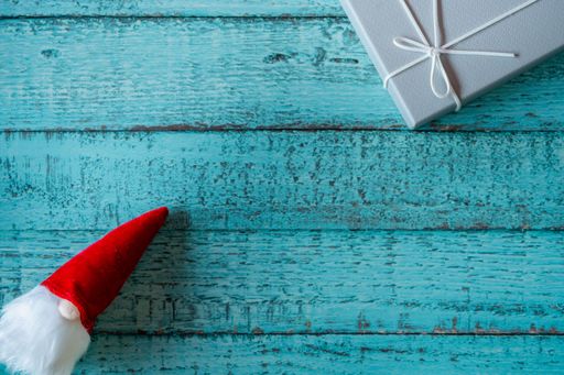 A top view of the Santa Claus toy with a long, white beard in a red hat near a gray gift box on a blue, wooden surface - the concept of New Year