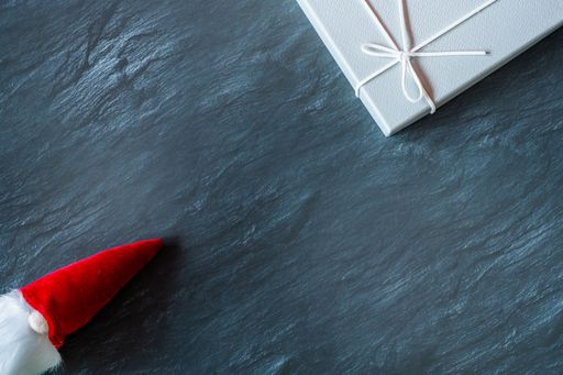 A top view of the Santa Claus toy with a long, white beard in a red hat near a gray gift box on a dark surface - the concept of New Year