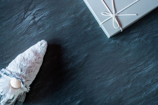 A top view of the gnome with a long, white beard and round nose in a gray hat near a gray gift box on a dark surface - the concept of New Year
