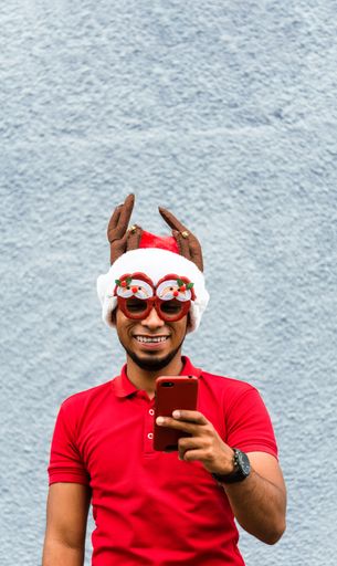 A vertical shot of a Latin man wearing a Christmas hat and glasses looking at his phone and laughing