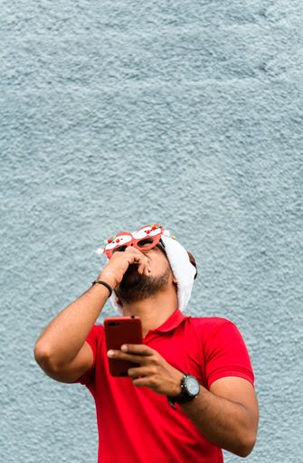 A vertical shot of a Latin man wearing a Christmas hat and glasses looking at his phone and laughing