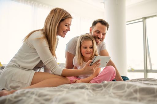 A happy young caucasian parents laughing while watching the tablet screen with their little daughter, on their bed in the bedroom
