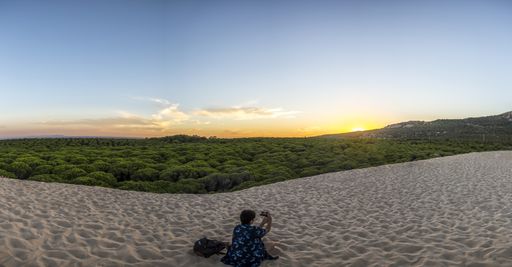 A back view of a woman sitting on the white sand and taking a photo of the beautiful sunset sky