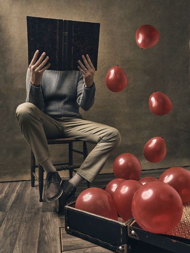 A vertical shot of a male reading a book and red balloons flying out of a vintage chest