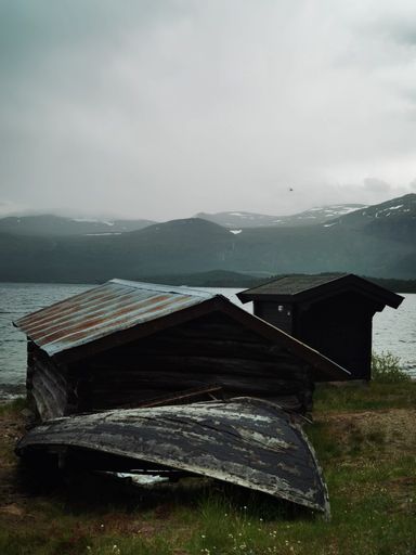 A vertical shot of dilapidated cabins on a seashore with mountains and a cloudy sky in the background