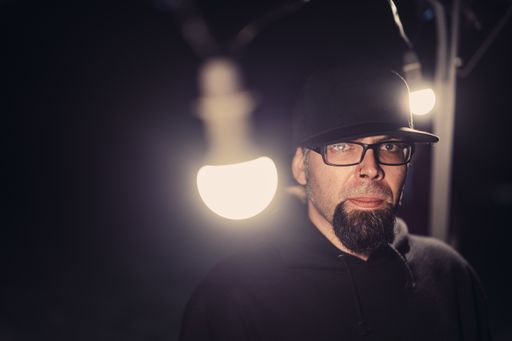 A closeup shot of a bearded man in black hoodie glasses and cap with lights