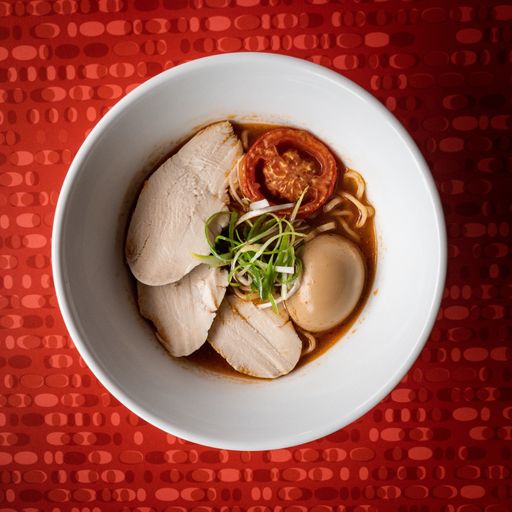A high angle shot of noodle soup with meat, tomato and an egg on a red surface