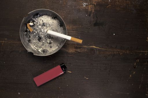 A top view of a lit cigarette in a dirty ashtray with a lighter on a wooden table