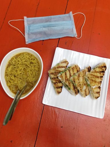 A vertical shot of noodle soup, tasty sandwiches, and a medical mask on a wooden surface - COVID-19