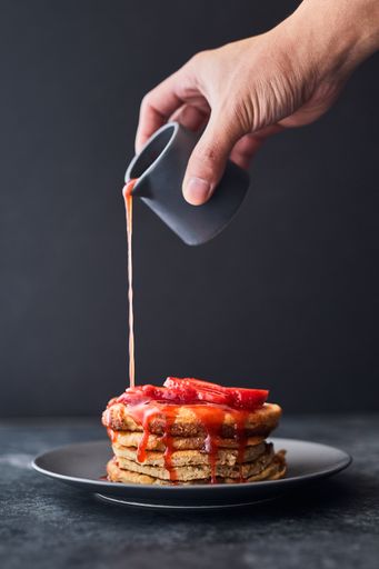 A vertical shot of a person pouring strawberry sauce on a stack of pancakes