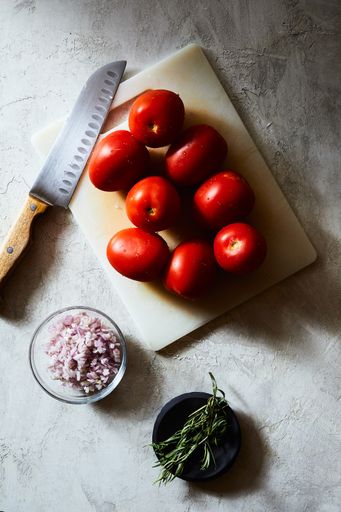 A vertical high angle shot of tomatoes on a white chopping board near a knife and chopped onions