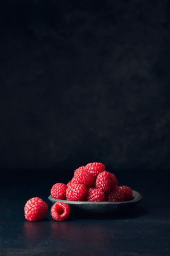 A vertical shot of raspberries in a plate on a black surface with a blurred background