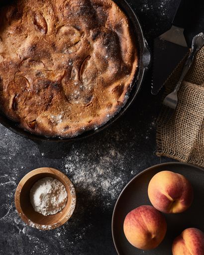 A vertical shot of a pie and a bowl of peaches on a black surface covered with flour