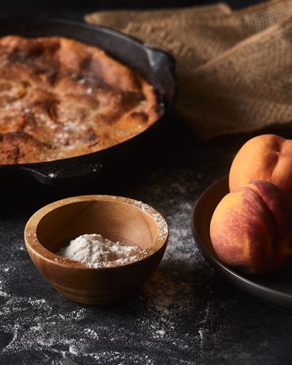 A vertical shot of a pie and a bowl of peaches on a black surface covered with flour
