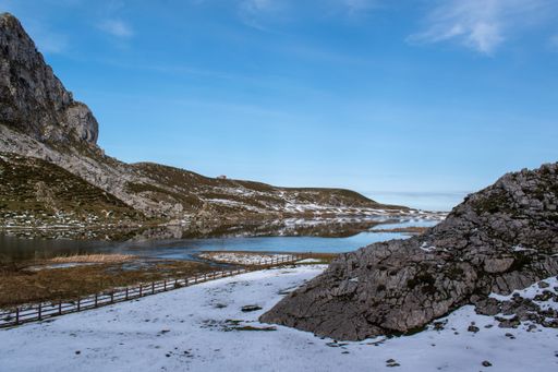 A scenic view of a lake against a fenced field covered with snow and a rocky mountain range