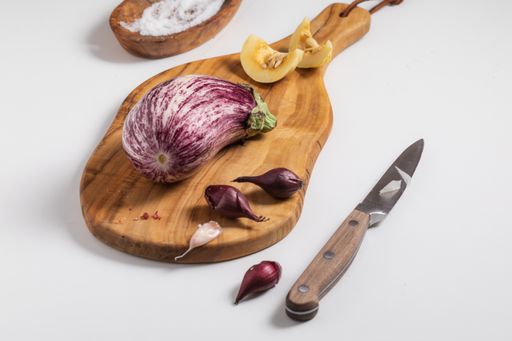 A closeup of nubia eggplant, garlic, and shallots on a small wooden cutting board, placed on a white surface with a  knife