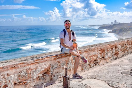An attractive sexy Hispanic male with sunglasses sitting on a parapet wall holding a water bottle with a wavy seascape behind