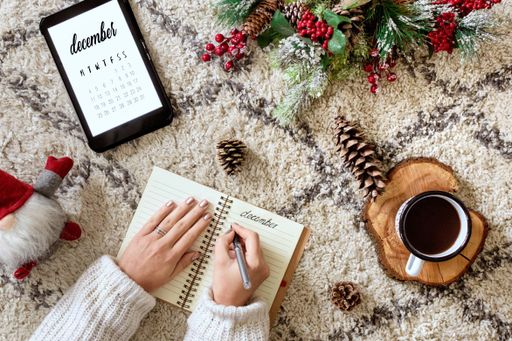 A top view of a caucasian woman writing in a planner next to a tablet showing the calendar, Christmas decorations, and a cup of coffee