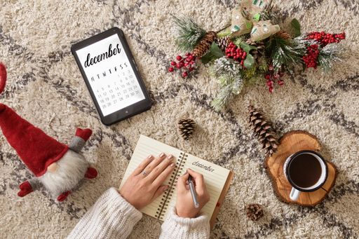 A top view of a caucasian woman writing in a planner next to a tablet showing the calendar, Christmas decorations, and a cup of coffee