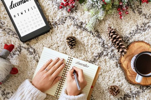 A top view of a caucasian woman writing in a planner next to a tablet showing the calendar, Christmas decorations, and a cup of coffee