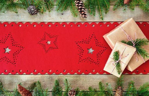 A top view of a wooden surface with a Christmas tablecloth, fir leaves and cones, and two wrapped gifts on it