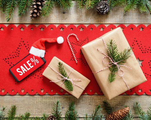 A top view of a wooden surface with a red Christmas table cloth, fir leaves, and cones, wrapped gifts, and a smartphone indicating Christmas sale