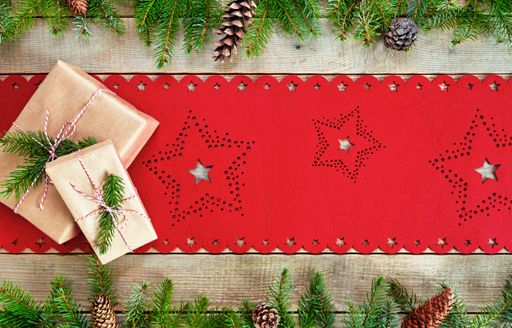 A top view of a wooden surface with a Christmas tablecloth, fir leaves and cones, and two wrapped gifts on it