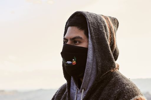 A young Hispanic man wearing a facemask and a hoodie, outdoors during the covid-19 pandemic
