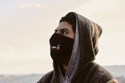 A young Hispanic man wearing a facemask and a hoodie, outdoors during the covid-19 pandemic