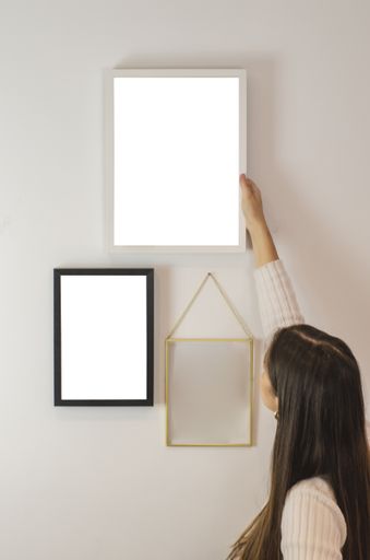 A vertical shot of a woman hanging an empty white-framed paper template on a wall