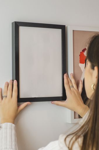 A vertical shot of a woman hanging an empty white-framed paper template on a wall