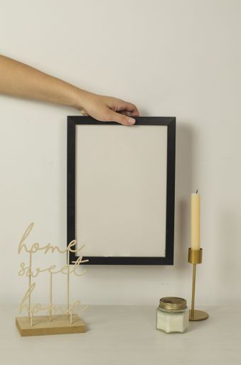 A hand putting a vertical black-framed poster mockup on a white background with decorative interior elements