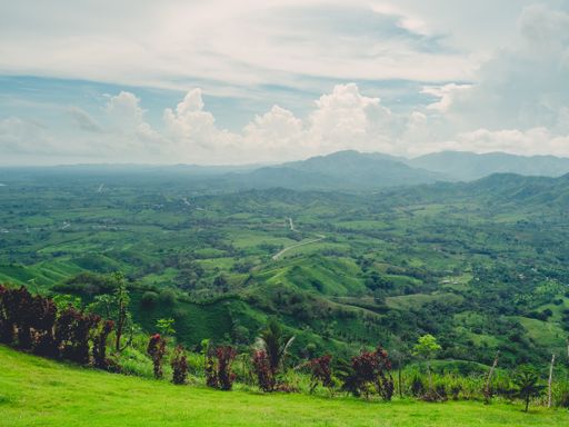 A landscape of hills covered in greenery with mountains on the background under a cloudy sky