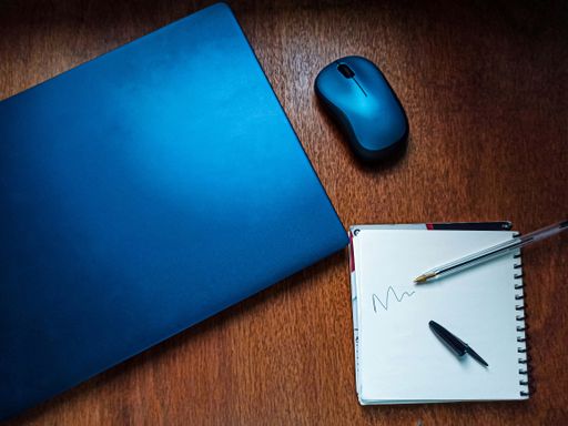 A top view shot of a pen, notebook, wireless mouse, and a laptop on a wooden surface