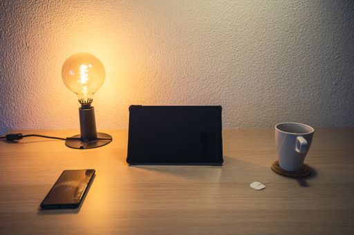 A white cup with tea on a wooden table with a lamp and a tablet on a blurred background