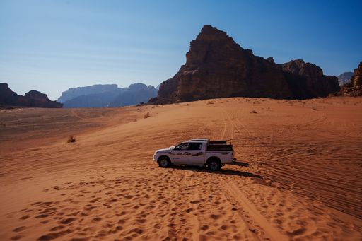 A white pickup truck in the middle of the sandy Wadi Rum desert in Jordan, with rock formations in the background on a sunny day