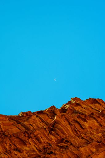 A vertical view of a steep sandstone mountain in the Wadi Rum desert with a new moon on blue sky background