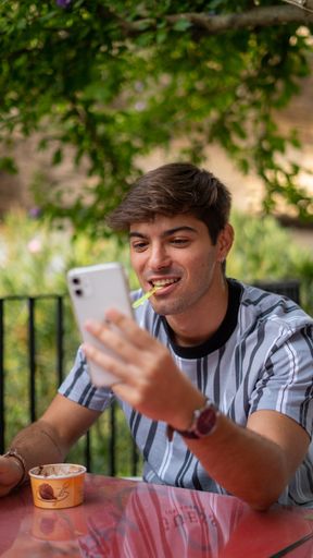 A vertical selective focus shot of a handsome young man eating ice cream at a cafe using his phone